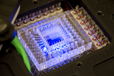 This 1 inch by 1 inch chip is part of the SETG instrument prototype. Tiny channels feed in the samples to be analyzed and control the fluidic circuitry on the chip. Blue light excites fluorescent dyes that help identify DNA within 3072 cubic chambers, each about the width of a human hair.