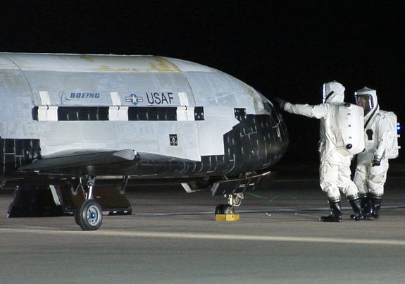 An X-37B robotic space plane sits on the Vandenberg Air Force base runway during post-landing operations on Dec. 3, 2010. Personnel in self-contained protective atmospheric suits conduct initial checks on the robot space vehicle after its landing. This same craft is due to launch again in fall 2012.