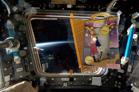 Arklu’s Stargazer Lottie doll, still in its box, floats in front of a window in the Cupola aboard the International Space Station.